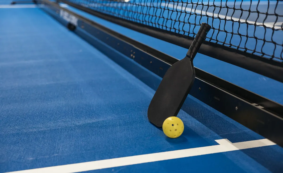 Raquette de pickleball et balle perforée posées au pied du filet sur un court bleu